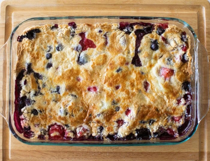 A rectangular glass baking dish holds a golden-brown cobbler with visible berries, likely blueberries and strawberries, peeking through the crust. The dish is placed on a wooden surface.