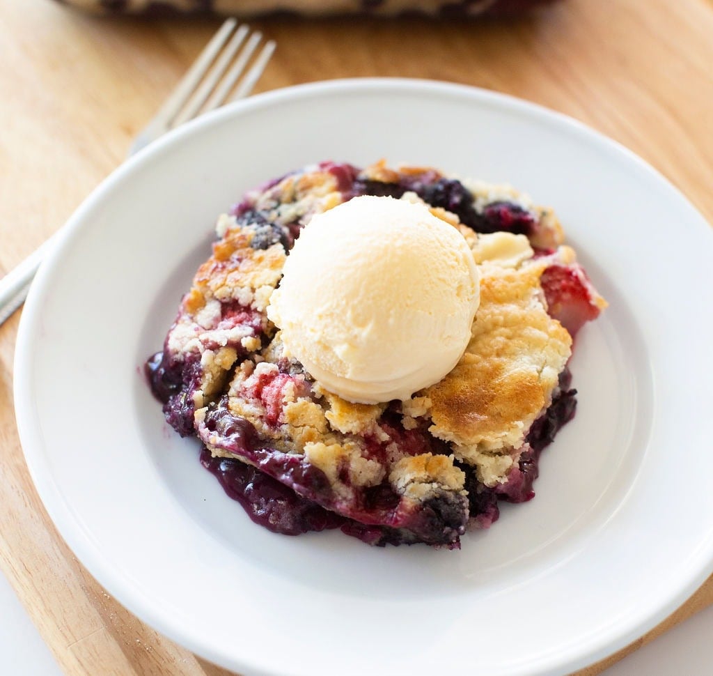 A serving of mixed berry crumble topped with a scoop of vanilla ice cream on a white plate. The dessert is set on a wooden surface with a fork and a hint of the larger dish in the background.