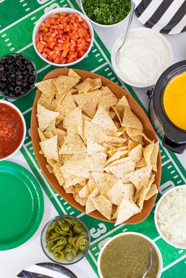 A football-themed tablecloth sets the scene for this fun nacho bar, with tortilla chips surrounded by bowls of salsa, cheese dip, tomatoes, olives, sour cream, jalapeños, onions, and fresh herbs.