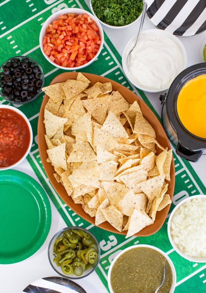 A football-themed tablecloth sets the scene for this fun nacho bar, with tortilla chips surrounded by bowls of salsa, cheese dip, tomatoes, olives, sour cream, jalapeños, onions, and fresh herbs.