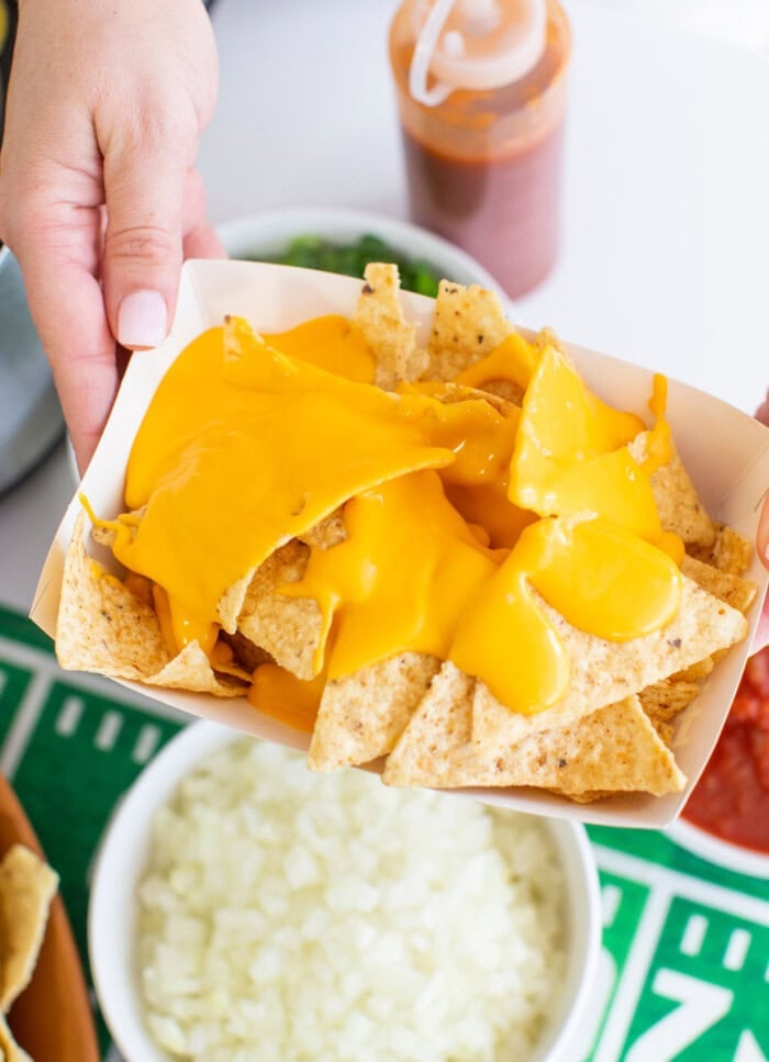 A hand holds a tray of nachos topped with melted cheese, with a bowl of chopped onions and a bottle of sauce nearby on a football-themed tablecloth for a nacho bar.