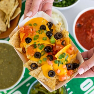 A person holds a tray of cheesy nachos topped with olives, tomatoes, and green onions, with bowls of salsa, green sauce, onions, and jalapeños nearby for a festive nacho bar.