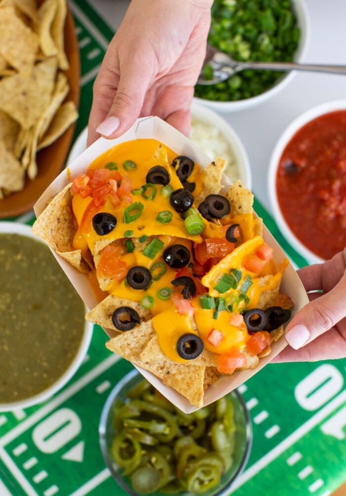 A person holds a tray of cheesy nachos topped with olives, tomatoes, and green onions, with bowls of salsa, green sauce, onions, and jalapeños nearby for a festive nacho bar.