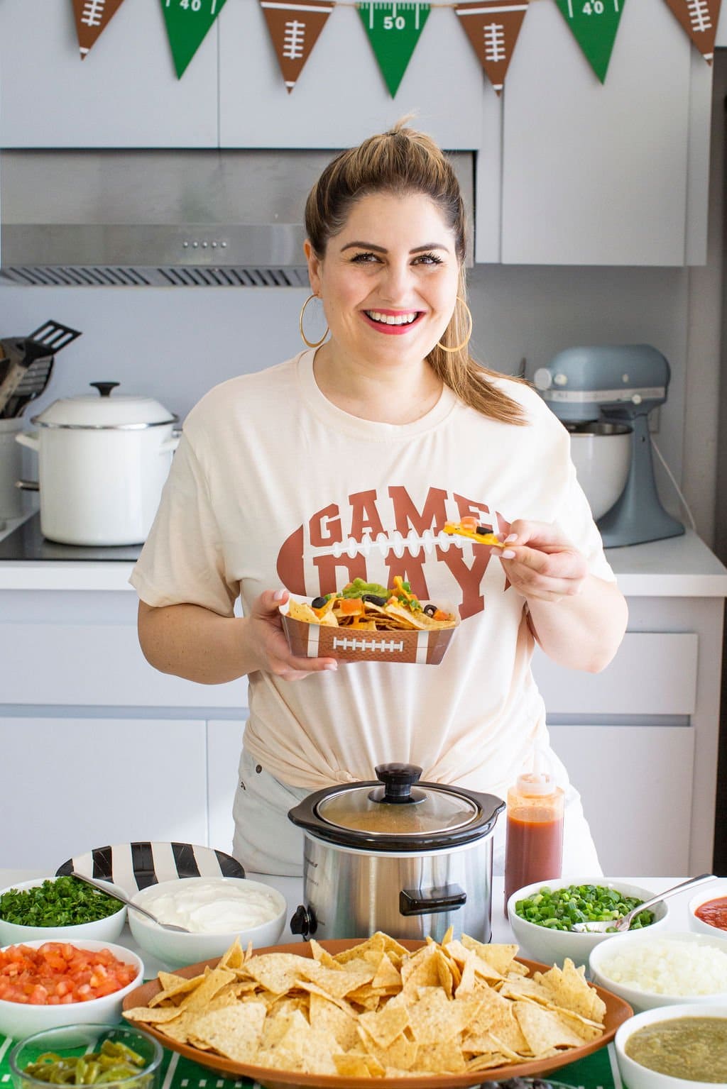 A person wearing a Game Day shirt holds a plate of nachos beside a festive nacho bar setup featuring assorted toppings, a small crockpot, hot sauce, and football-themed decorations.