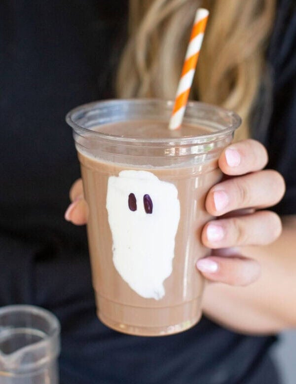 A person holding a clear plastic cup of chocolate milk with a white ghost decoration and an orange-striped straw. The persons nails are painted light pink.