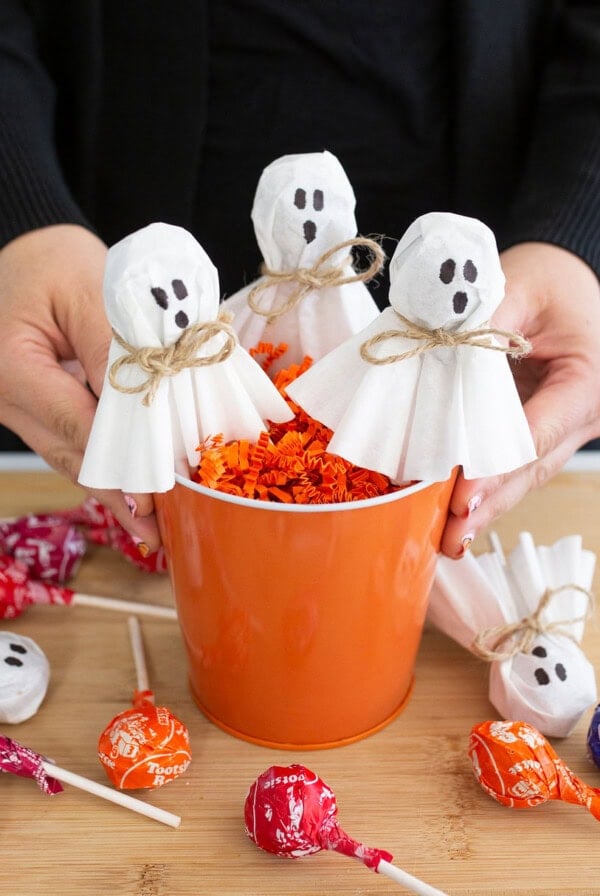 A person holds an orange bucket filled with orange paper shreds and ghost-themed lollipops. The lollipops have white tissue covers, drawn faces, and twine ties, surrounded by more colorful lollipops on a wooden surface.