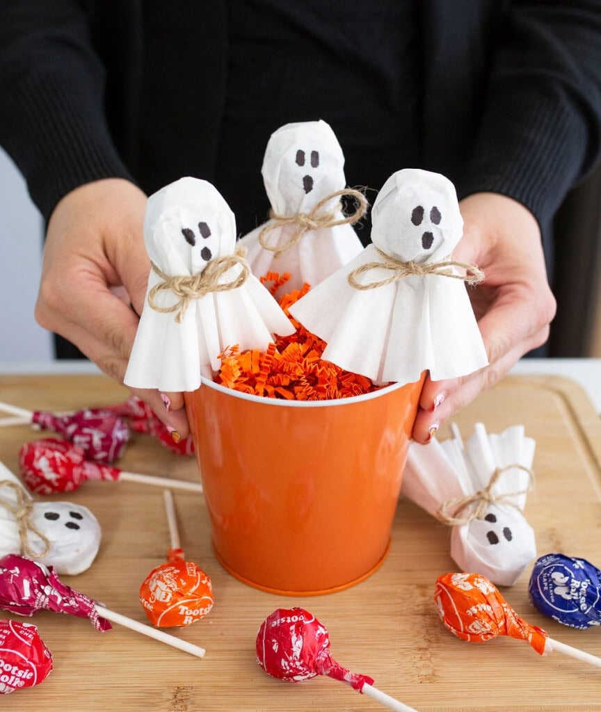 A person holds an orange bucket filled with orange paper shreds and ghost-themed lollipops. The lollipops have white tissue covers, drawn faces, and twine ties, surrounded by more colorful lollipops on a wooden surface.