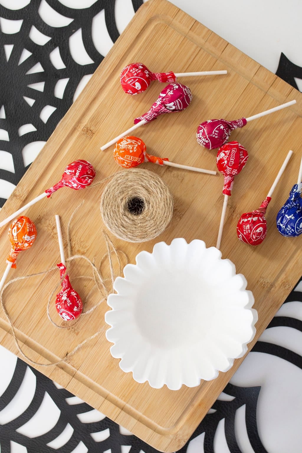 Halloween candy lollipops and twine sit on a wooden board beside a white scalloped plate, with a black-and-white spiderweb background.