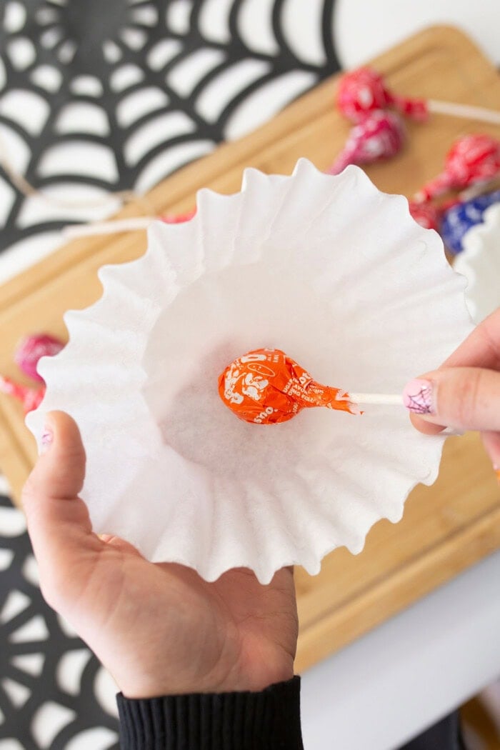 Hands wrap a coffee filter around a lollipop to create a Halloween candy treat, with more lollipops and a decorative spiderweb mat on the wooden surface.