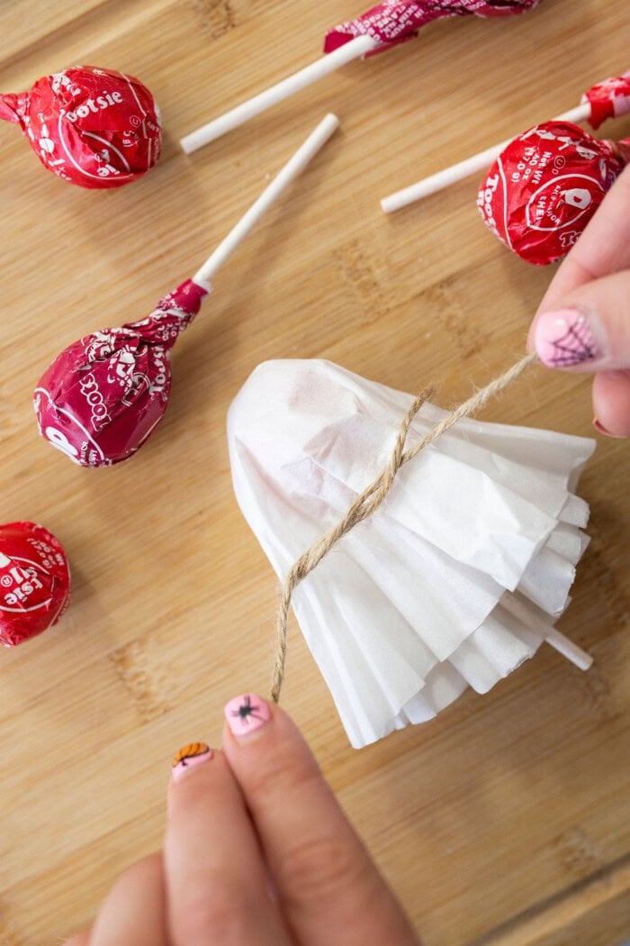 Hands wrap a white coffee filter around a red lollipop and tie it with twine to make a ghost, with more Halloween candy on a wooden surface.
