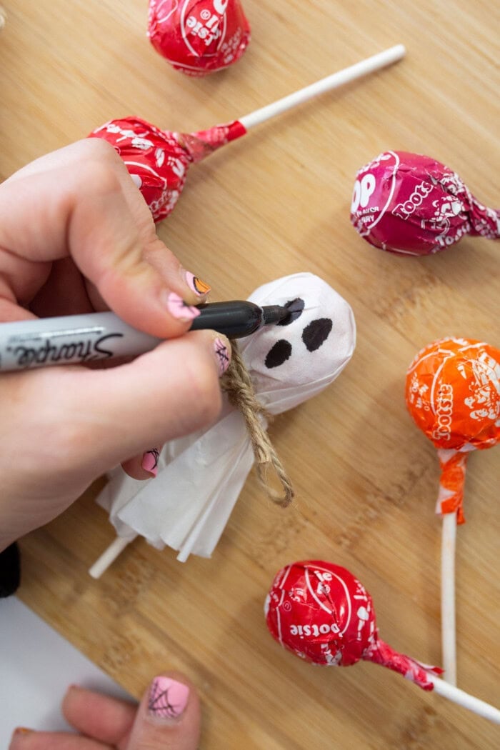 A person draws a ghost face on a tissue-covered lollipop with a black marker, while other ghost lollipops sit on a wooden surface.