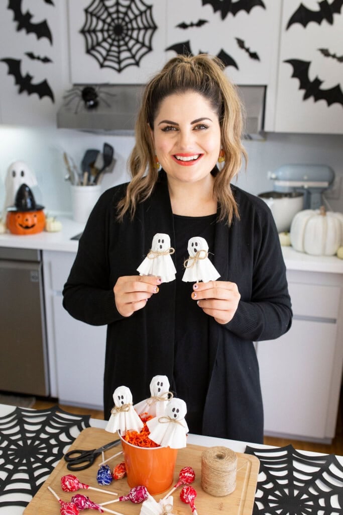 A person smiles while holding handmade ghost decorations, likely for Halloween. The kitchen background features spider and bat decor. A table displays more ghosts, twine, and lollipops in an orange container.