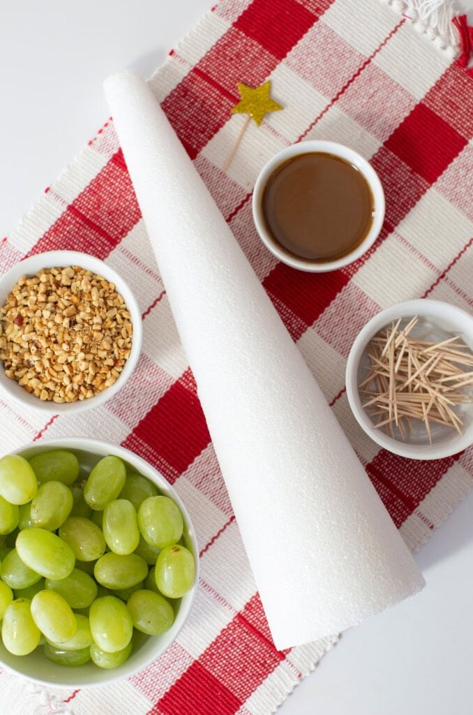 A white cone, green grapes, chopped nuts, toothpicks, caramel sauce, and a gold star are arranged on a red and white checkered cloth to create a festive fruit Christmas tree—perfect as a whimsical Christmas dessert.