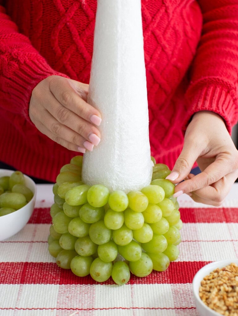 A person in a red sweater attaches green grapes onto a white foam cone, crafting a festive fruit Christmas tree. Bowls of grapes and chopped nuts rest on a red-and-white checkered tablecloth, perfect for an eye-catching Christmas dessert.