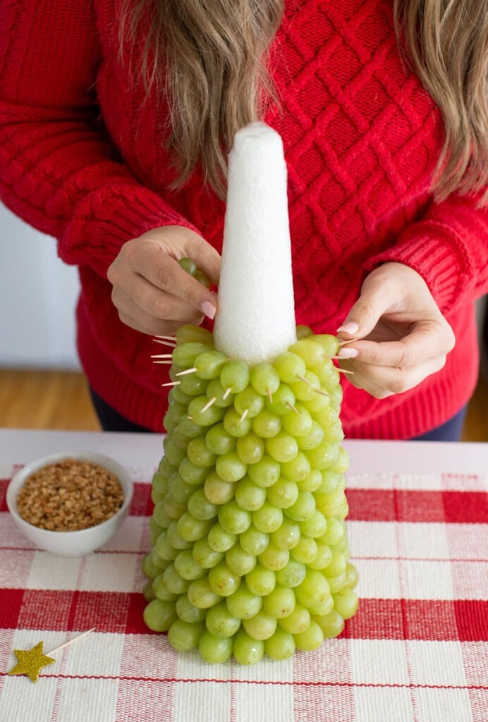 A person in a red sweater attaches green grapes to a white cone with toothpicks, crafting a festive fruit Christmas tree, with a bowl of nuts on the table nearby for the perfect Christmas dessert.