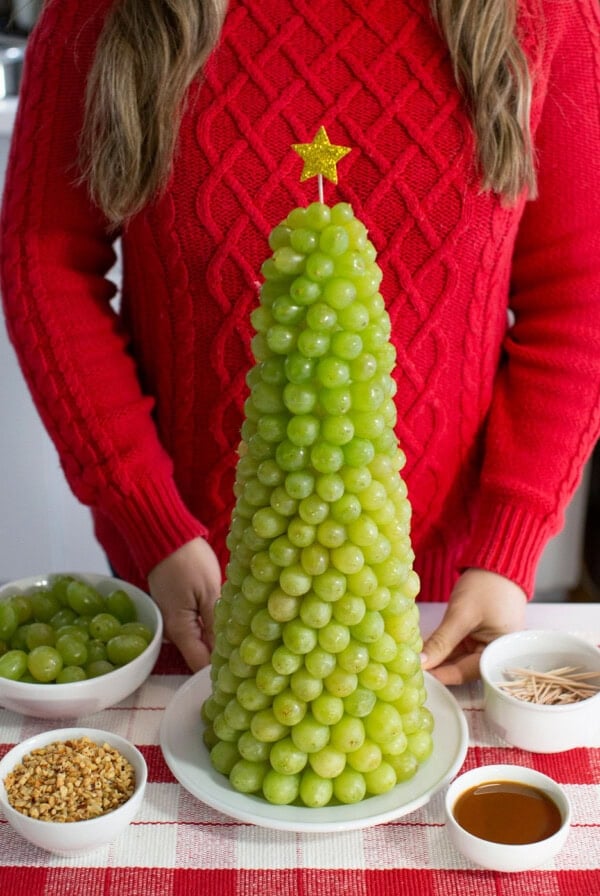 A person in a red sweater stands behind a tall fruit Christmas tree made of green grapes, shaped like a Christmas tree and topped with a yellow star—perfect as a festive Christmas dessert. Bowls of grapes, nuts, toothpicks, and caramel sauce are nearby.