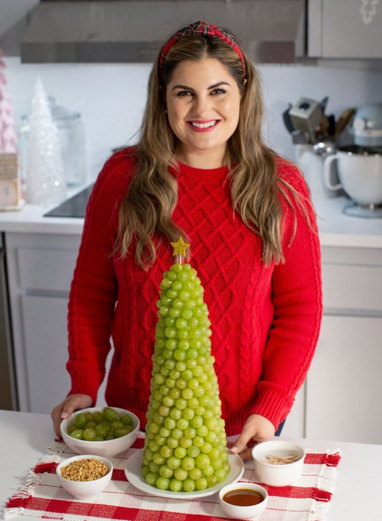 A woman in a red sweater stands in a kitchen, smiling behind a tall, cone-shaped fruit Christmas tree made of green grapes. Bowls of grapes, nuts, syrup, and slivered almonds are arranged on the counter for this festive Christmas dessert.