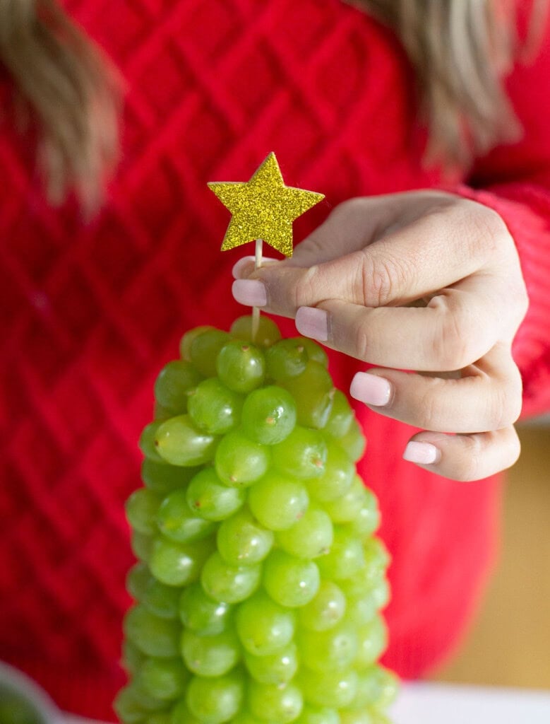 A hand places a glittery gold star on top of a fruit Christmas tree made of green grapes. The person, wearing a red textured sweater, adds the final touch to this festive christmas dessert.