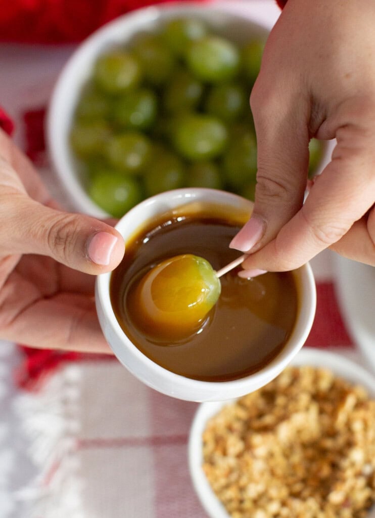 A person holds a green grape on a toothpick, dipping it into caramel sauce—perfect for a festive christmas dessert—with bowls of grapes and crushed nuts in the background, ideal for building a fruit christmas tree.