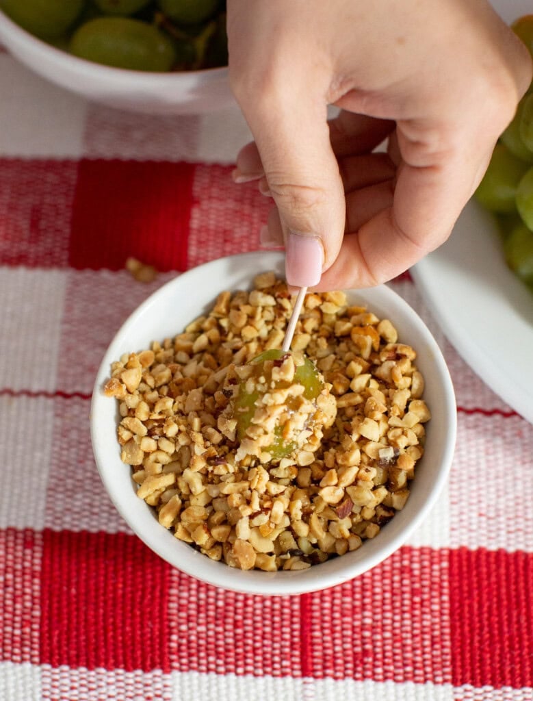 A hand dips a grape with a toothpick into a bowl of crushed nuts on a red and white checkered tablecloth, creating the perfect bite for a festive fruit Christmas tree or easy Christmas dessert. Nearby sit a plate of grapes and another bowl.