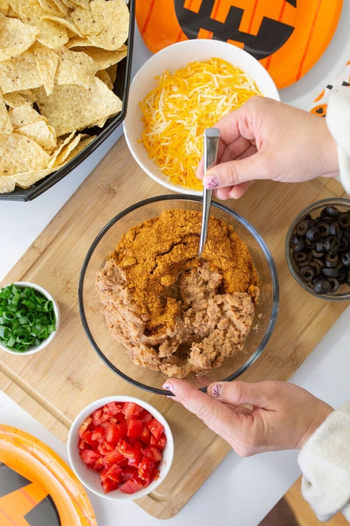 Hands mixing refried beans and taco seasoning in a bowl on a wooden board. Nearby are bowls with shredded cheese, chopped tomatoes, sliced black olives, and chopped green onions. A tray of tortilla chips is beside them on a festive orange plate.