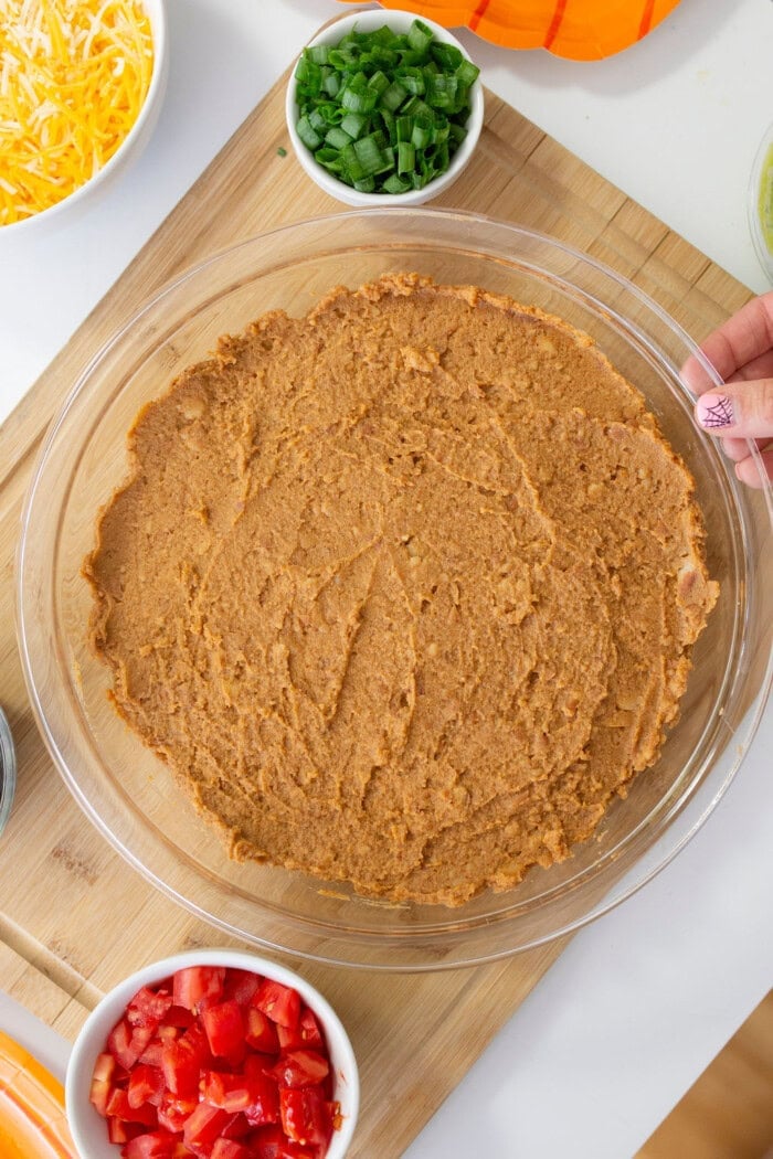 A glass pie dish filled with refried beans for a bean dip, with fresh toppings nearby.