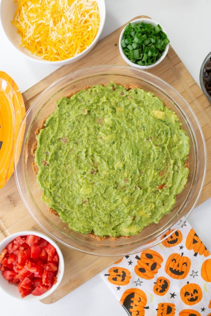 A glass dish filled with guacamole stands on a wooden board. Nearby bowls hold shredded cheese, chopped tomatoes, and sliced green onions. A Halloween-themed napkin featuring jack-o-lanterns is placed beside the setup.