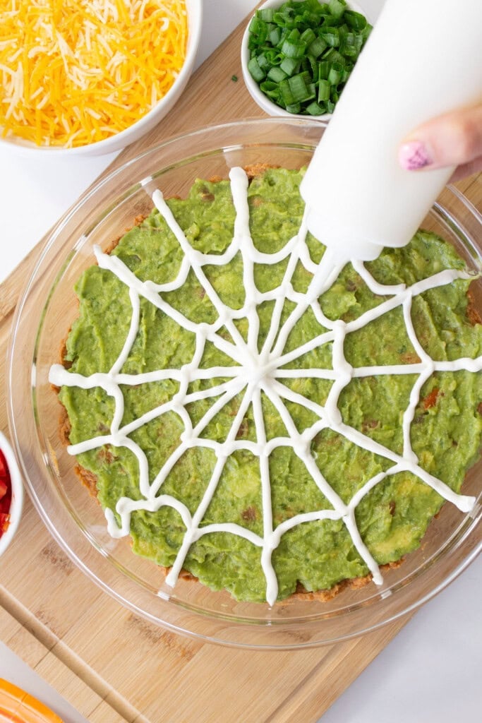 A hand decorates guacamole in a glass dish with a sour cream spiderweb pattern. Surrounding the dish are bowls of shredded cheese, chopped green onions, and diced tomatoes.