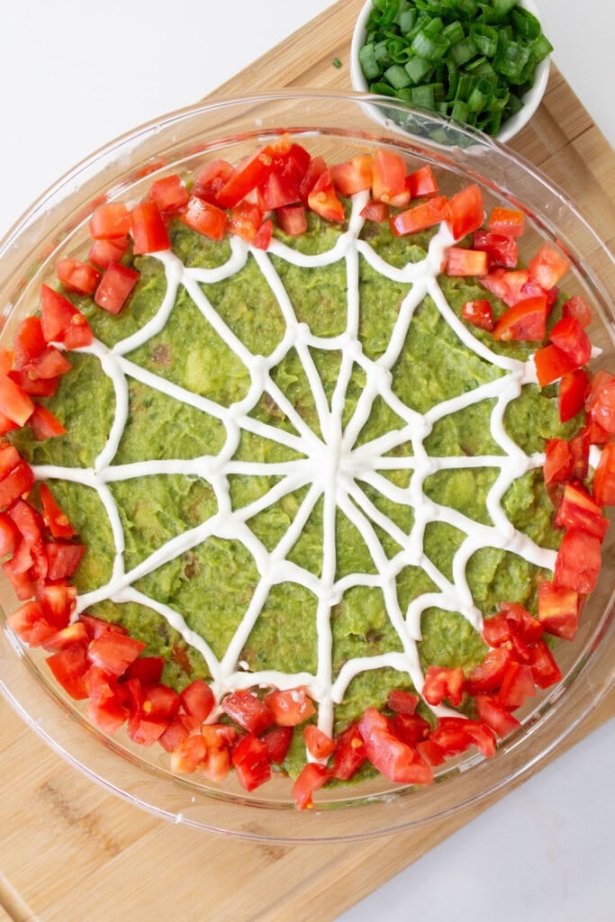 A round platter of guacamole features a spiderweb design made from sour cream. The dip is bordered by chopped tomatoes, and a small bowl of chopped green onions is in the background, all placed on a wooden surface.