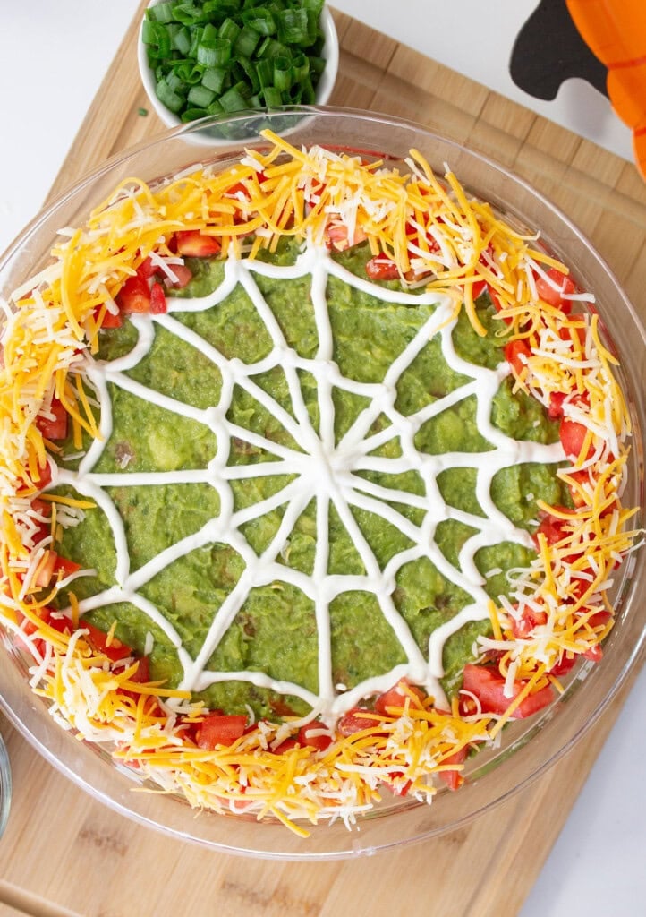 A layered dip in a round dish features a top layer of guacamole decorated with a sour cream spiderweb design. The edges are garnished with shredded cheese and diced tomatoes. A small bowl of chopped green onions is nearby on a wooden board.