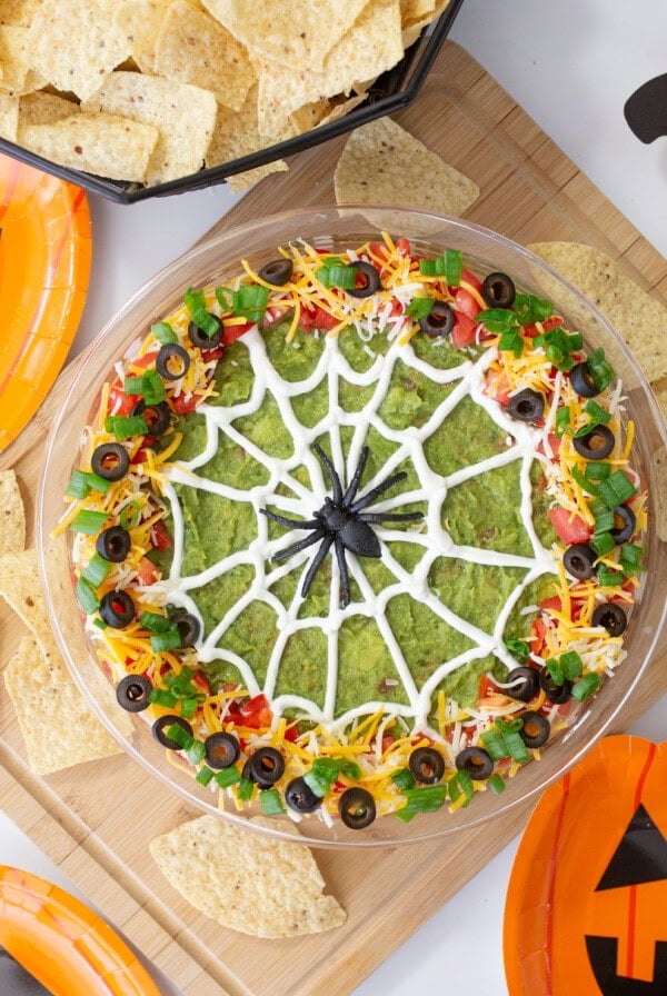 A festive guacamole dip styled with a spider web pattern made of sour cream, surrounded by toppings like shredded cheese, tomatoes, olives, and green onions. Its part of a Halloween-themed setup with tortilla chips on the side.