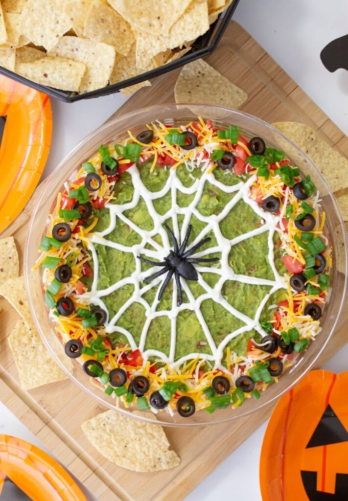 A festive guacamole dip styled with a spider web pattern made of sour cream, surrounded by toppings like shredded cheese, tomatoes, olives, and green onions. Its part of a Halloween-themed setup with tortilla chips on the side.
