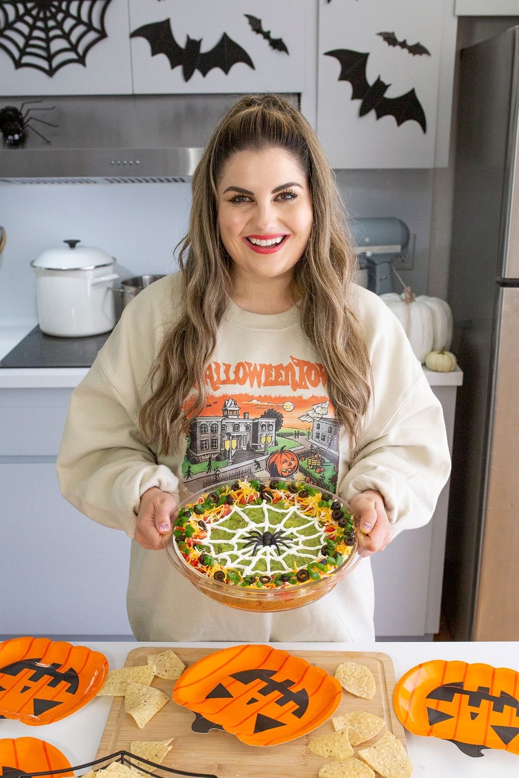 A person with long hair, wearing a Halloween-themed sweater, holds a festive pie with a spider web design. The kitchen is decorated with black bats and spider webs. Orange plates with Jack-o-lantern designs are on the counter nearby.