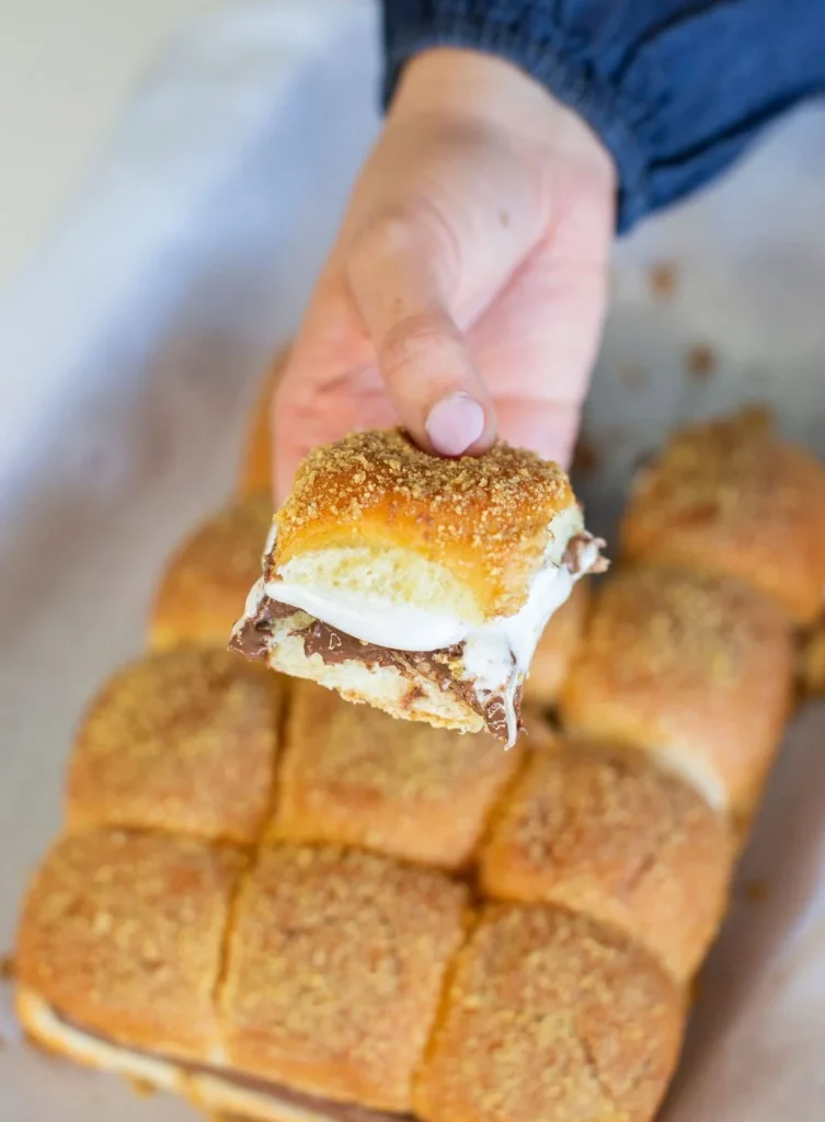 A hand holds a pull-apart piece of smores bread, showing the melted chocolate, marshmallow, and crumbled topping. Below, a tray with more golden-brown pull-apart smores bread is visible in the background.