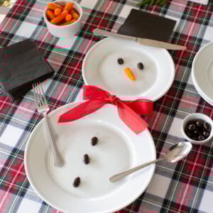 Two plates arranged like a snowman on a plaid tablecloth. One plate has a carrot nose and olive eyes, the other has olives for buttons. A red ribbon is the scarf. Utensils mimic arms. Nearby are black napkins and small bowls of carrots and olives.