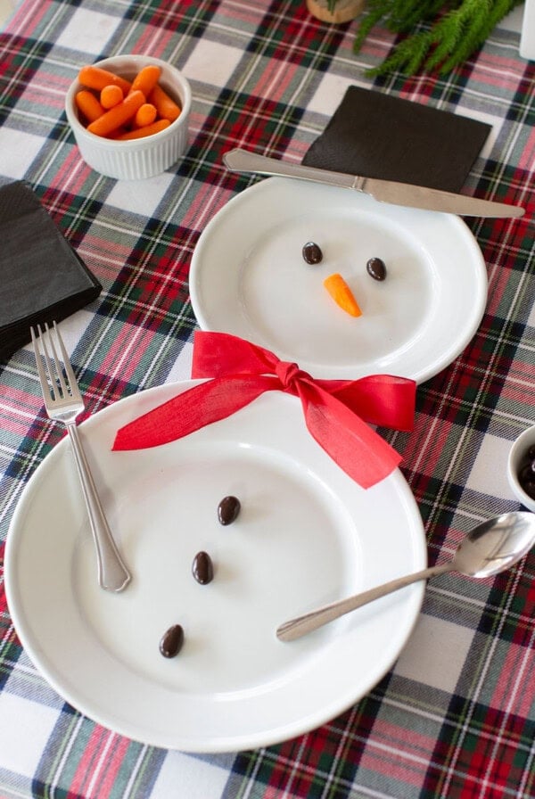 Two plates arranged like a snowman on a plaid tablecloth. One plate has a carrot nose and olive eyes, the other has olives for buttons. A red ribbon is the scarf. Utensils mimic arms. Nearby are black napkins and small bowls of carrots and olives.