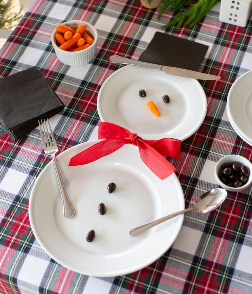 Two plates arranged like a snowman on a plaid tablecloth. One plate has a carrot nose and olive eyes, the other has olives for buttons. A red ribbon is the scarf. Utensils mimic arms. Nearby are black napkins and small bowls of carrots and olives.