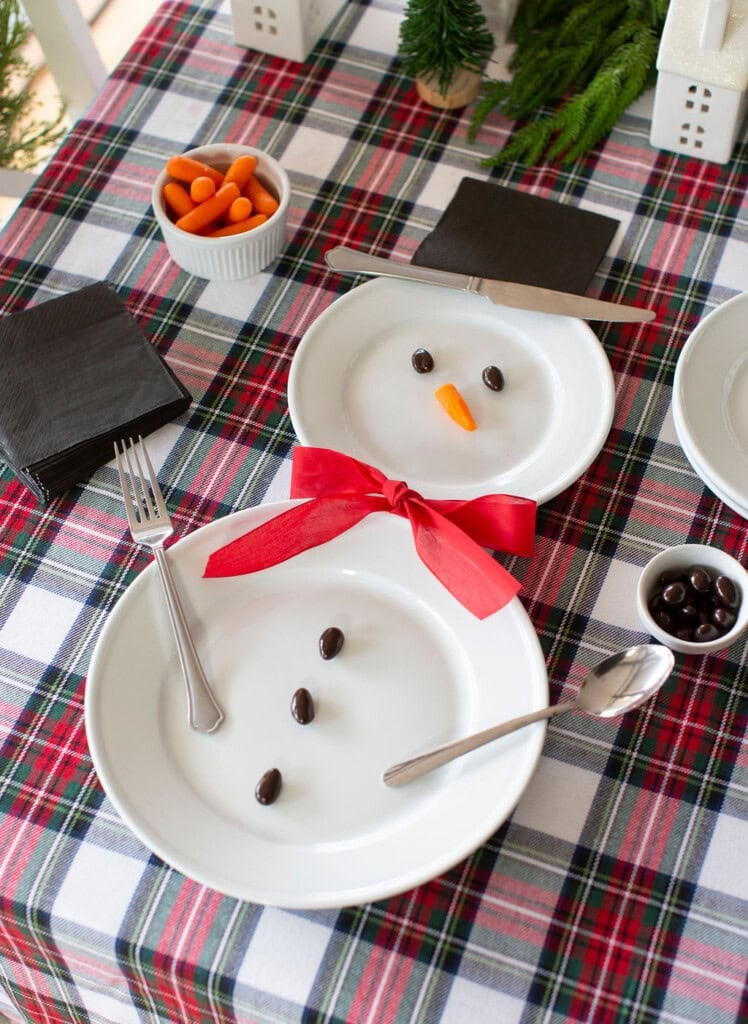 Festive table setting with plates arranged to resemble a snowman. A red ribbon forms a bow tie, a carrot is the nose, and dark candies are eyes and buttons. Utensils serve as arms. A plaid tablecloth and small bowls of carrots and candies complete the scene.