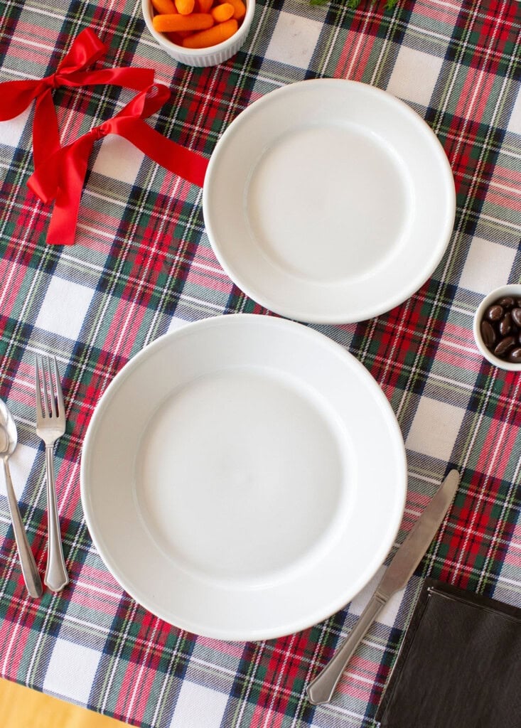 A table is set with two white plates on a red and green plaid tablecloth. Silverware and a black napkin are on the left, with bowls of baby carrots, chocolates, and a red ribbon nearby.