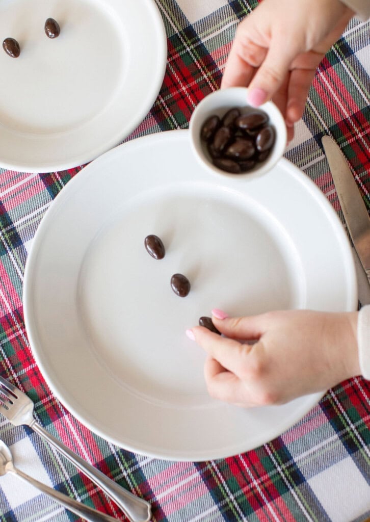 Hands placing chocolate-covered almonds onto a white plate. A small bowl with more almonds is visible. The table is covered with a red, green, and white plaid tablecloth. Utensils are nearby.