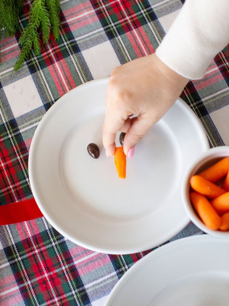 A hand places a small carrot on a white plate next to an olive. The plate is on a red and green plaid tablecloth. A bowl with more small carrots is nearby.