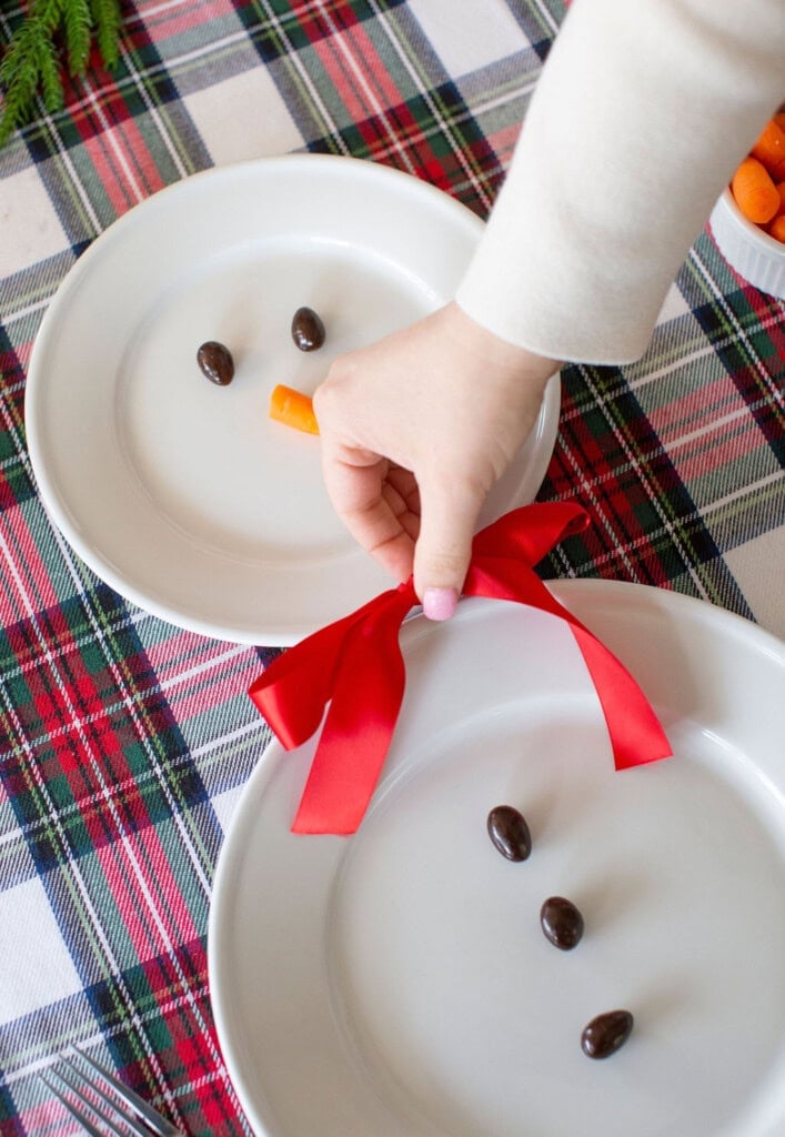 A hand places a carrot on a white plate with chocolate candies, forming a snowman face, on a plaid tablecloth. Another plate has candies with a red bow below it, suggesting a snowman with a scarf.