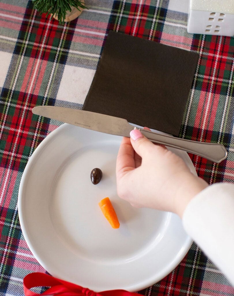 A hand places a butter knife on a white plate with a carrot piece and a black olive, arranged like a face on a plaid tablecloth. A black napkin and part of a white small house decoration are visible in the background.