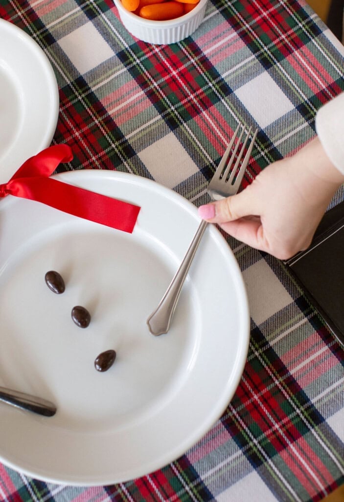 A hand adjusts a fork on a plaid tablecloth with three chocolate-covered almonds on a white plate tied with a red ribbon. A bowl of sliced carrots is visible in the background.