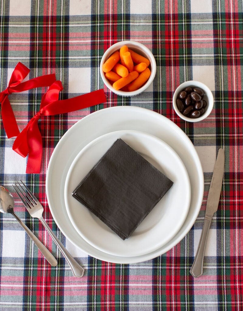 A table setting on a red, white, and green plaid cloth with a red ribbon. There are stacked white plates, a black napkin, silver utensils, and two bowls containing orange carrot slices and chocolate-covered items.