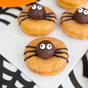 Halloween donuts topped with chocolate spider bodies, candy eyes, and black icing legs, arranged on a white rectangular plate for a cute and festive treat.