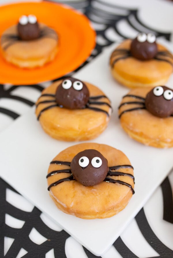 Four glazed Halloween donuts are decorated with chocolate spider bodies, candy eyes, and black icing legs. One sits on an orange plate, while the others are arranged on a white serving board over a spiderweb-patterned tablecloth.