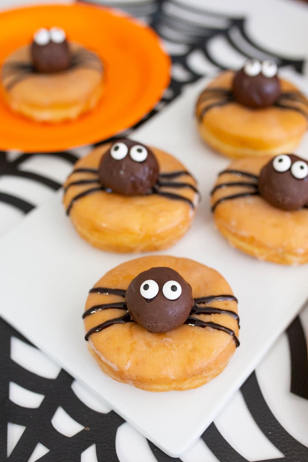 Four glazed Halloween donuts are decorated with chocolate spider bodies, candy eyes, and black icing legs. One sits on an orange plate, while the others are arranged on a white serving board over a spiderweb-patterned tablecloth.