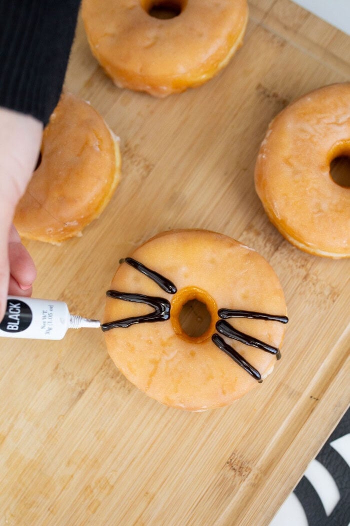 A glazed donut is decorated with black icing to create stripes across the top, with more glazed donuts in the background. This Halloween donuts idea is simple and festive.