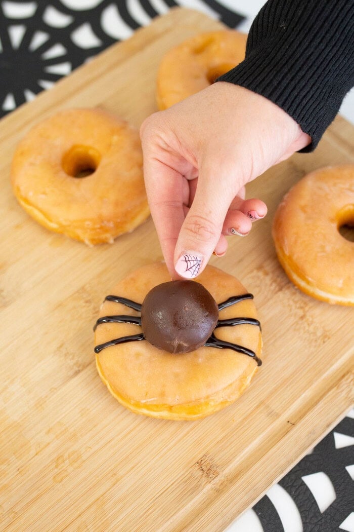 A hand places a chocolate cake pop onto a glazed donut decorated with black icing legs to create a spider shape. More glazed donuts sit on a wooden board nearby, making these Halloween donuts a fun and festive treat.